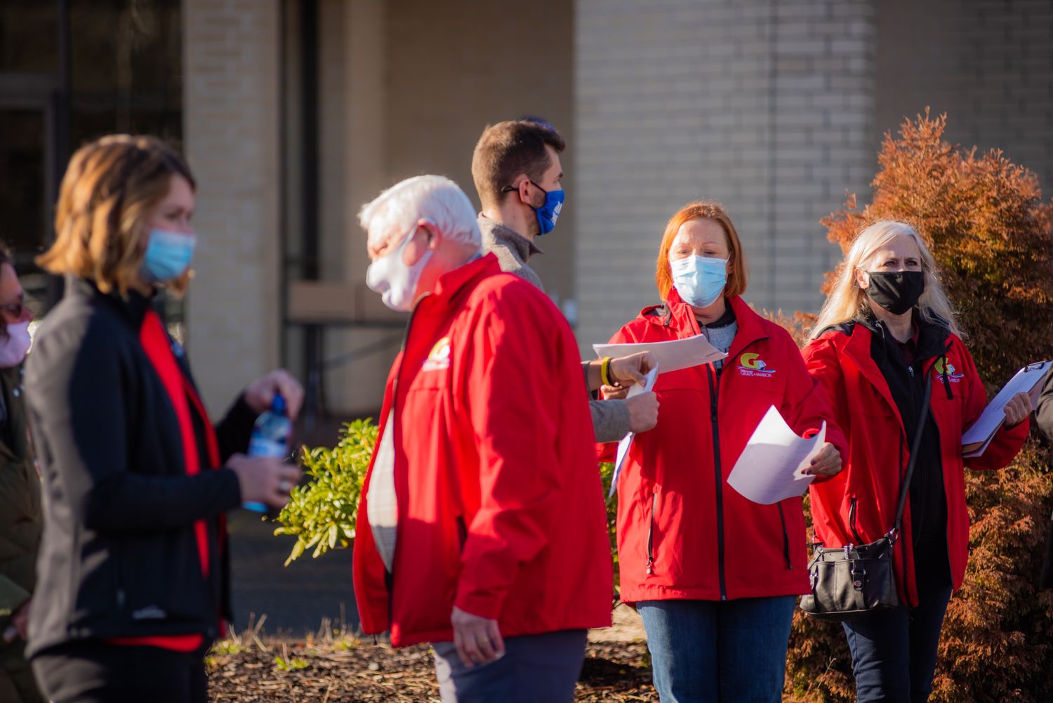Four people in red jackets and face masks stand outdoors holding papers while a woman in a dark jacket holds a water bottle.