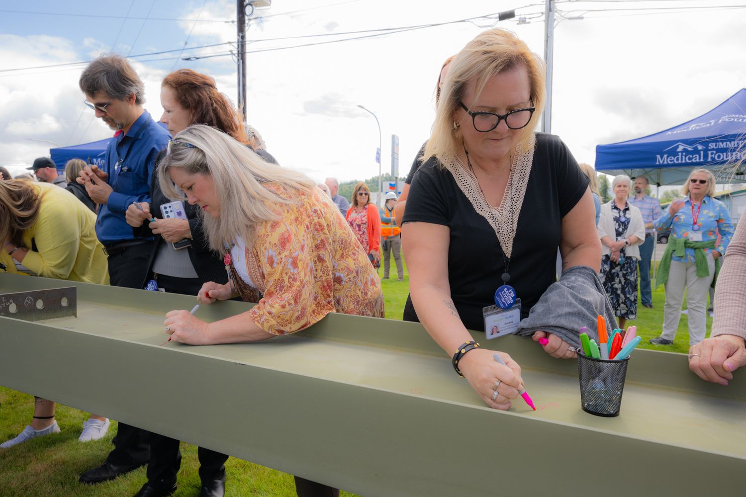 Group of people gather around a green steel beam at an outdoor event, signing it with colorful markers while a woman in glasses smiles.
