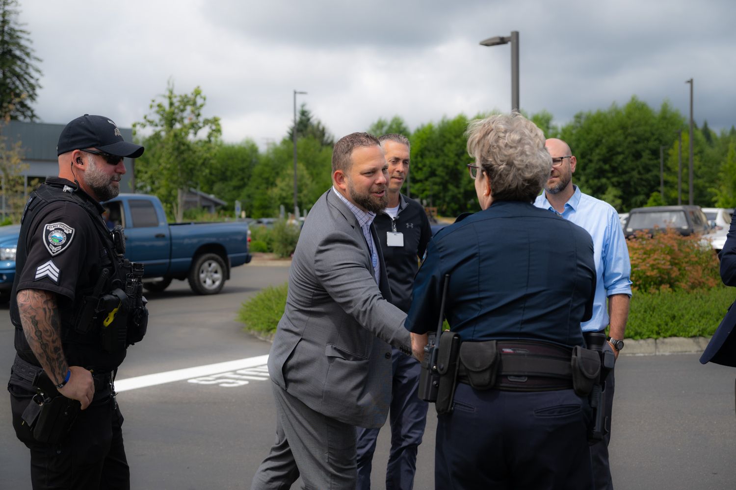 A man in a gray suit shakes hands with a uniformed police officer in a parking lot as another officer and two men look on.