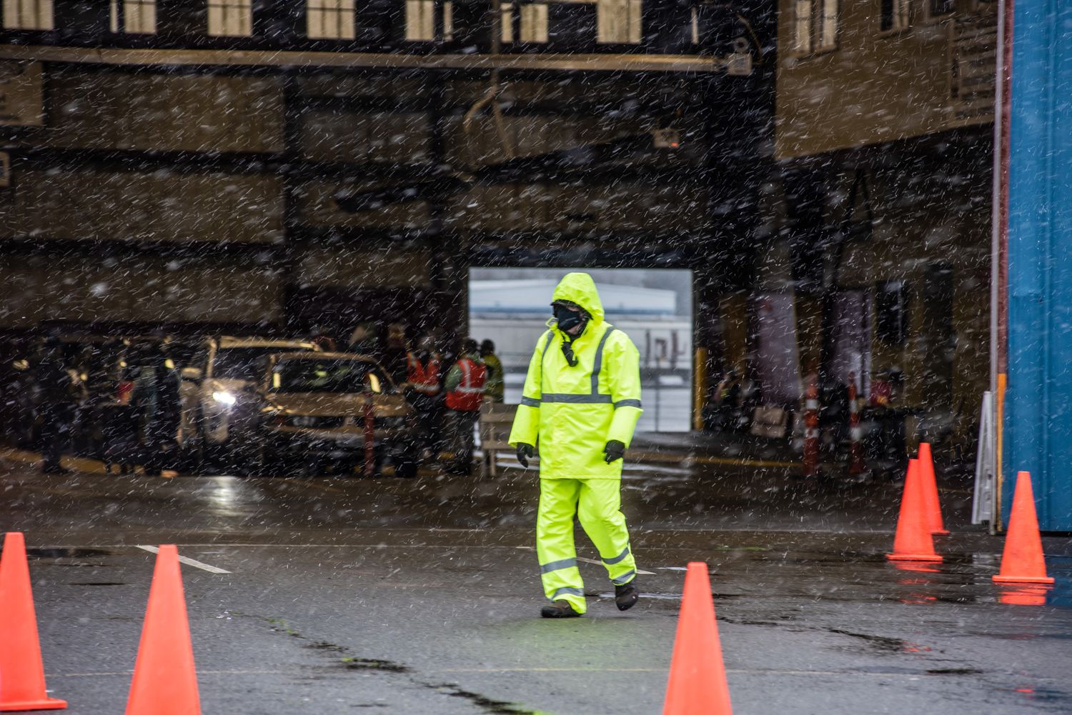 Worker in a high-visibility yellow rain suit and mask walks past orange traffic cones on a wet street during a snowstorm.