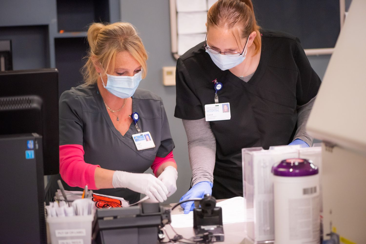 Two healthcare workers in scrubs, face masks and gloves handle medical supplies and equipment at a clinical workstation in a lab.