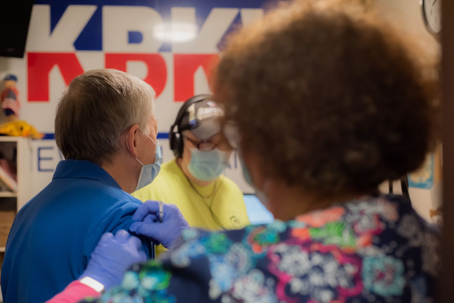 A masked health worker in blue gloves administers a vaccine to a masked man at a KBPK event while another worker in a headset looks on.