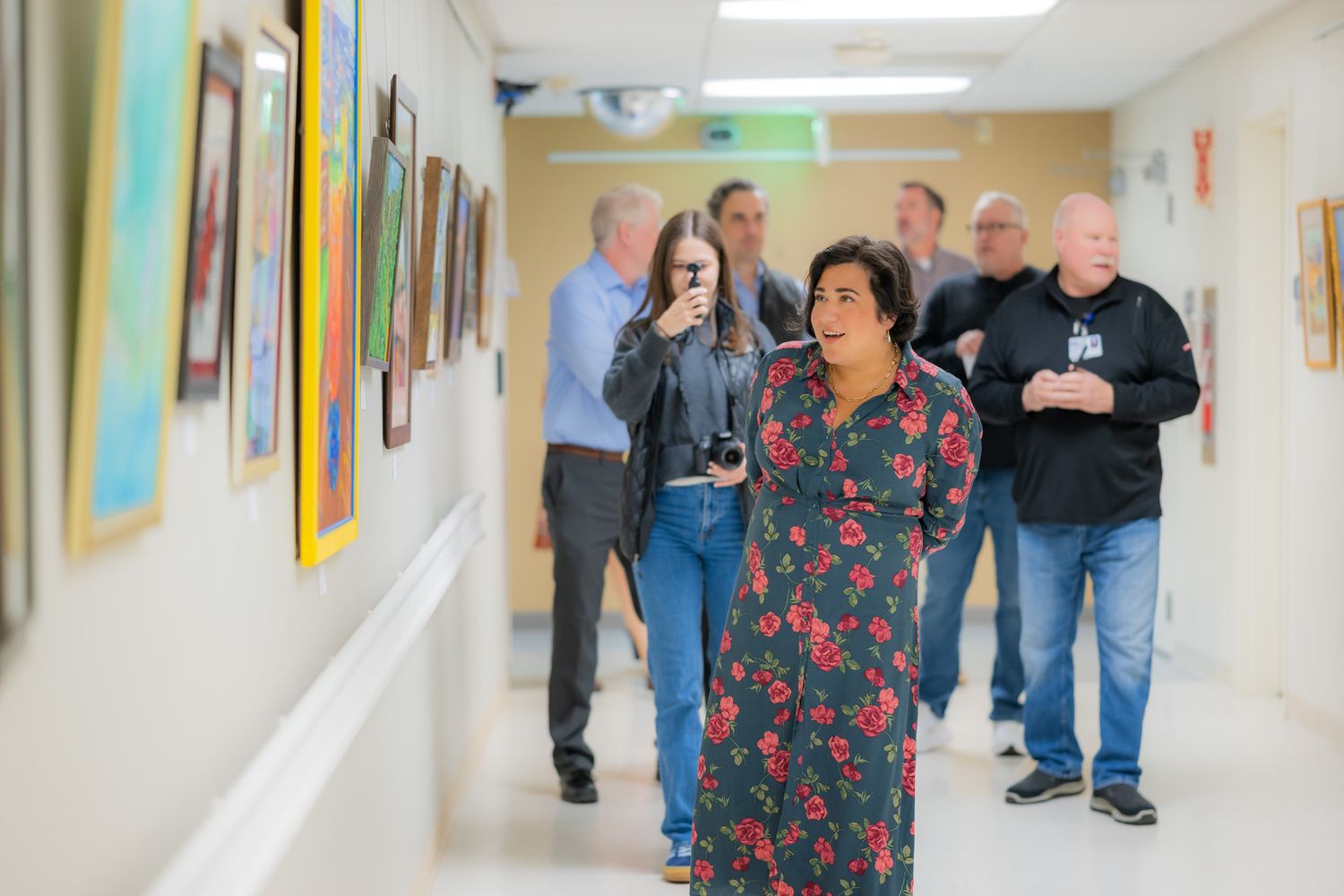 Visitors walk down a bright gallery hallway lined with colorful framed paintings while one woman in a floral dress smiles.