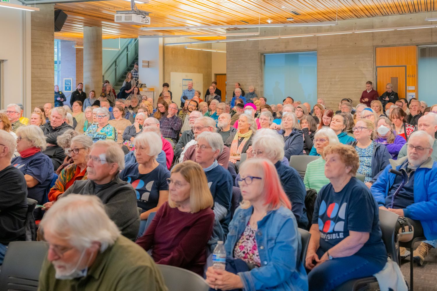 Large audience of mostly older adults seated in a community hall, attentively listening to a speaker during a public meeting.