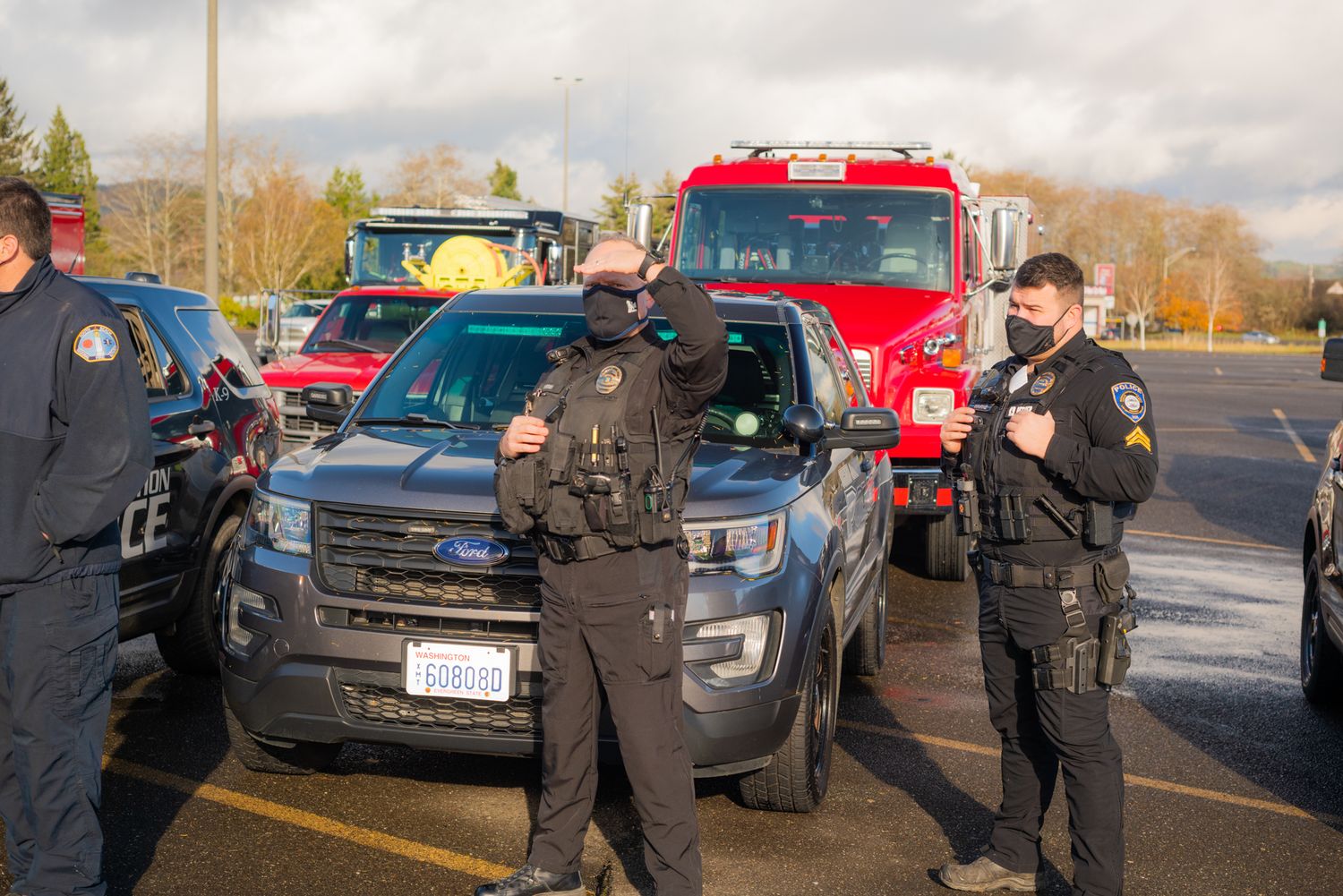 Two masked police officers in tactical vests stand in front of a patrol SUV with a red fire truck parked behind them.