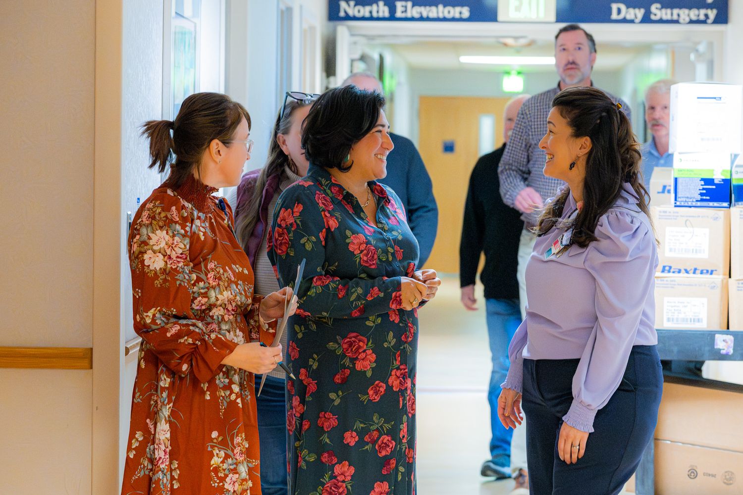 Three women in floral and lavender outfits chat in a bright hospital hallway near the North Elevators and Day Surgery sign.