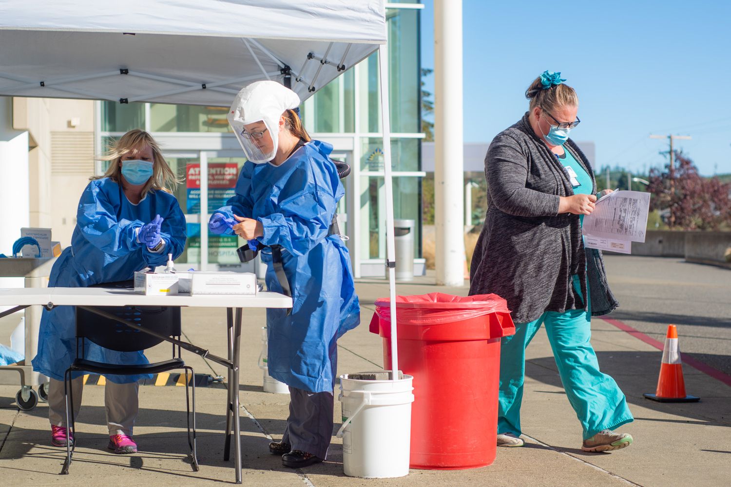 Healthcare workers in blue protective gowns and face shields staff an outdoor tent testing station as another person walks past.