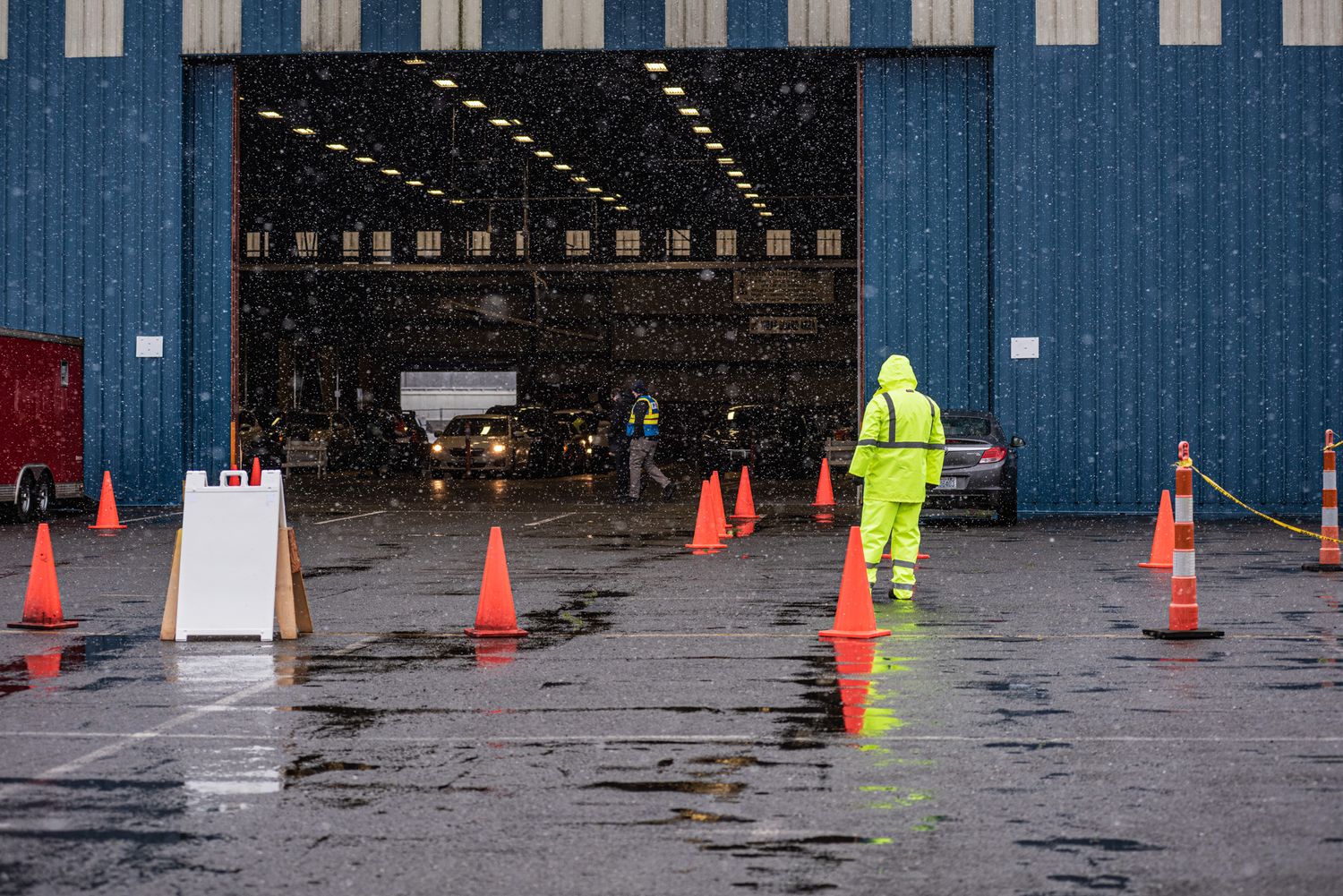 A worker in a bright yellow rain jacket stands among orange traffic cones on wet pavement outside a large warehouse entrance.