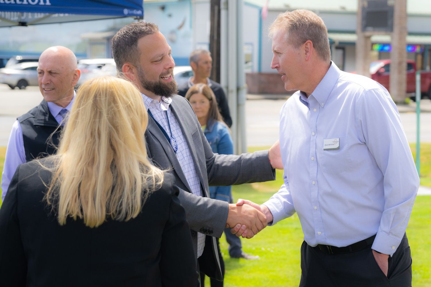 Two men in business attire shake hands outdoors on a sunny day while a blonde woman and other onlookers stand nearby.