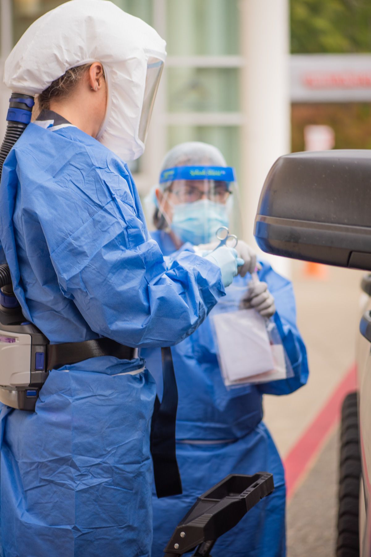 Healthcare workers in blue protective gowns, face shields and a powered respirator hood conduct drive-through testing beside a vehicle.
