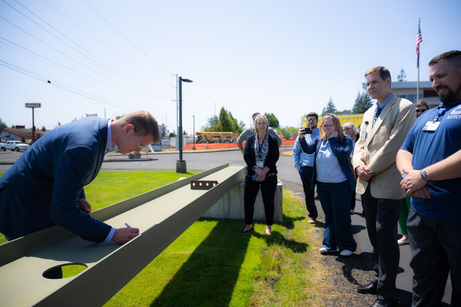 A man in a blue suit leans over a concrete structure outdoors while a group of onlookers watches during a ceremony on a sunny day.