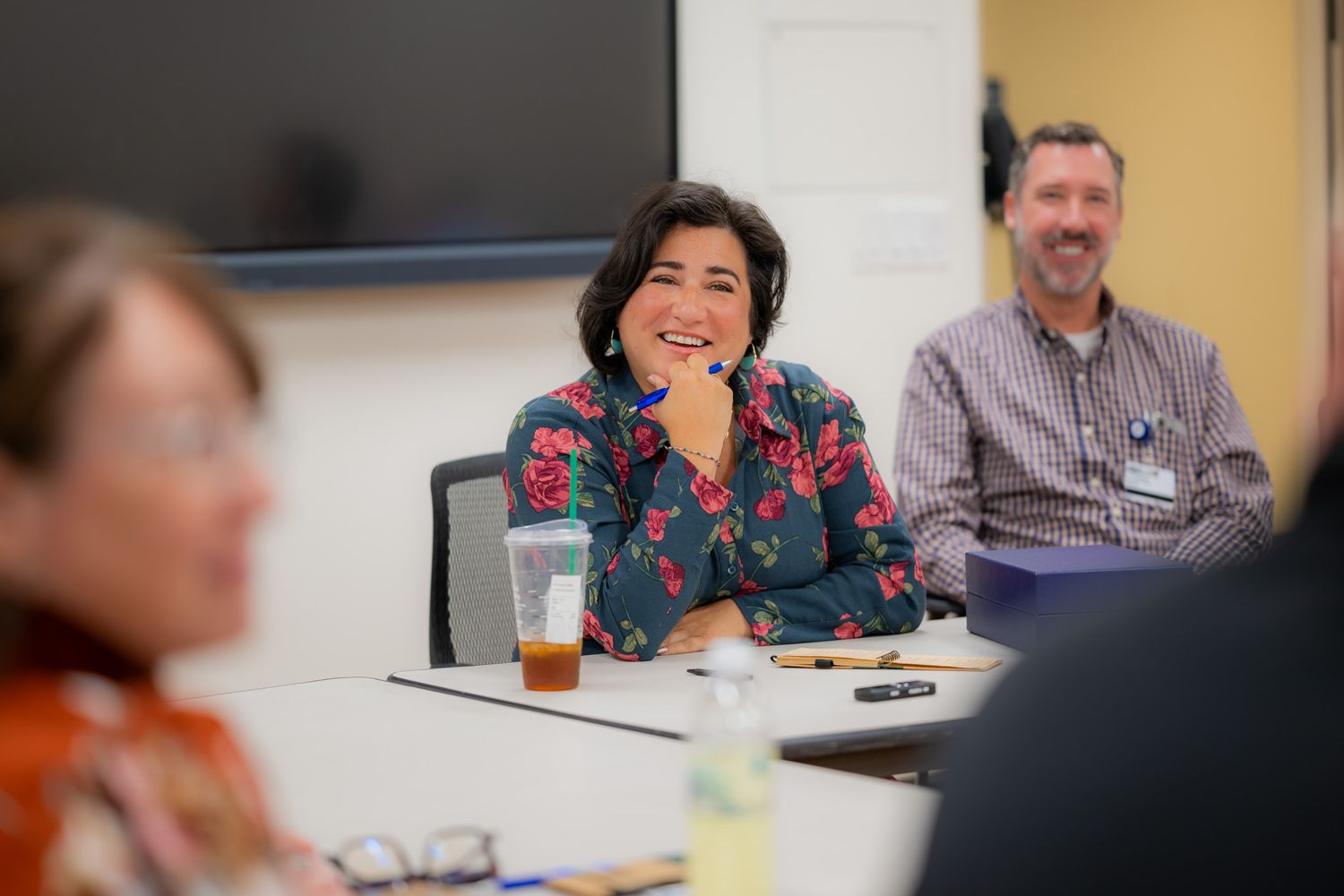 Smiling woman in a floral blouse rests her chin on her hand at a conference table while a man in a plaid shirt smiles behind her.