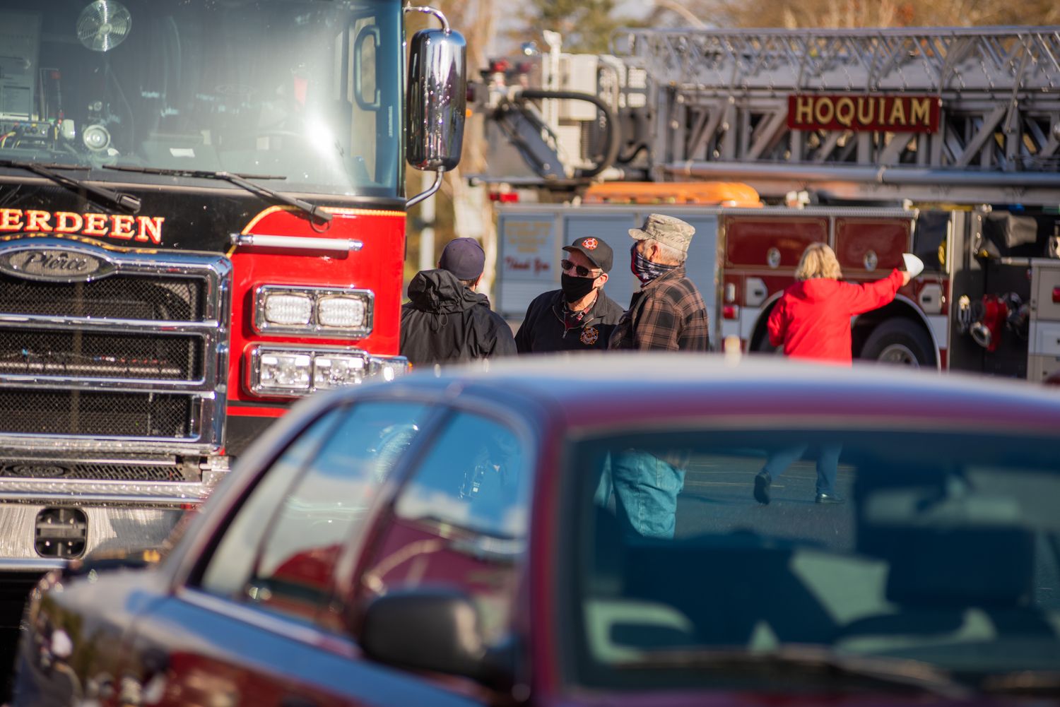 Firefighters in masks stand beside a red fire engine and ladder truck, seen through the windshield of a parked car.