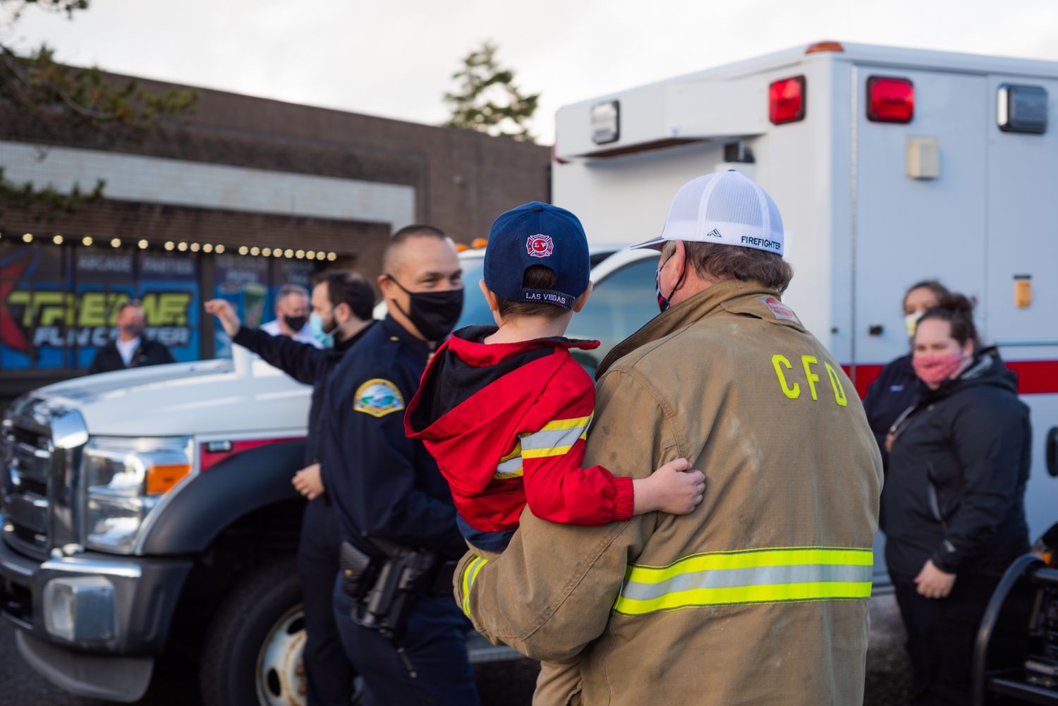 Masked firefighter in a tan CFD jacket holds a child in a red jacket and blue cap beside an ambulance and fire truck as onlookers gather.