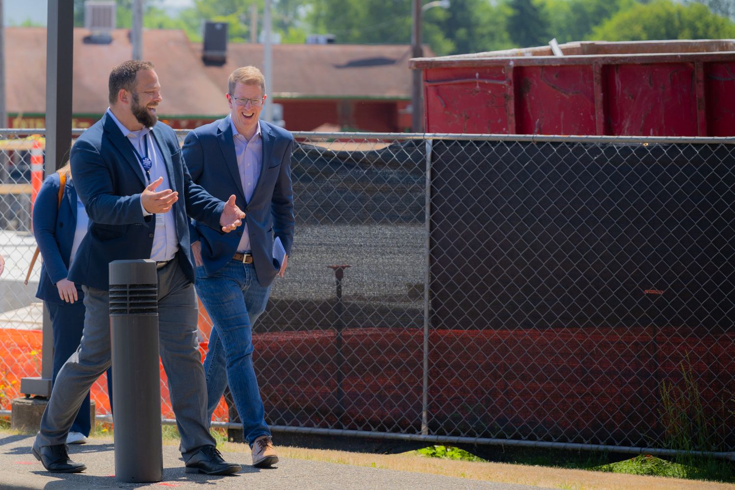 Two men in business casual attire walk and talk outdoors beside a chain-link fence with red construction barriers in the background.