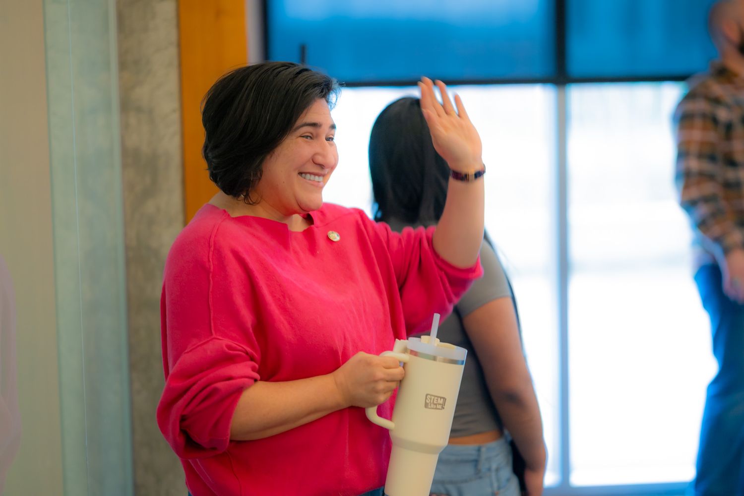 Smiling woman in a bright pink sweater waves while holding a white tumbler with a straw, standing indoors near large windows.