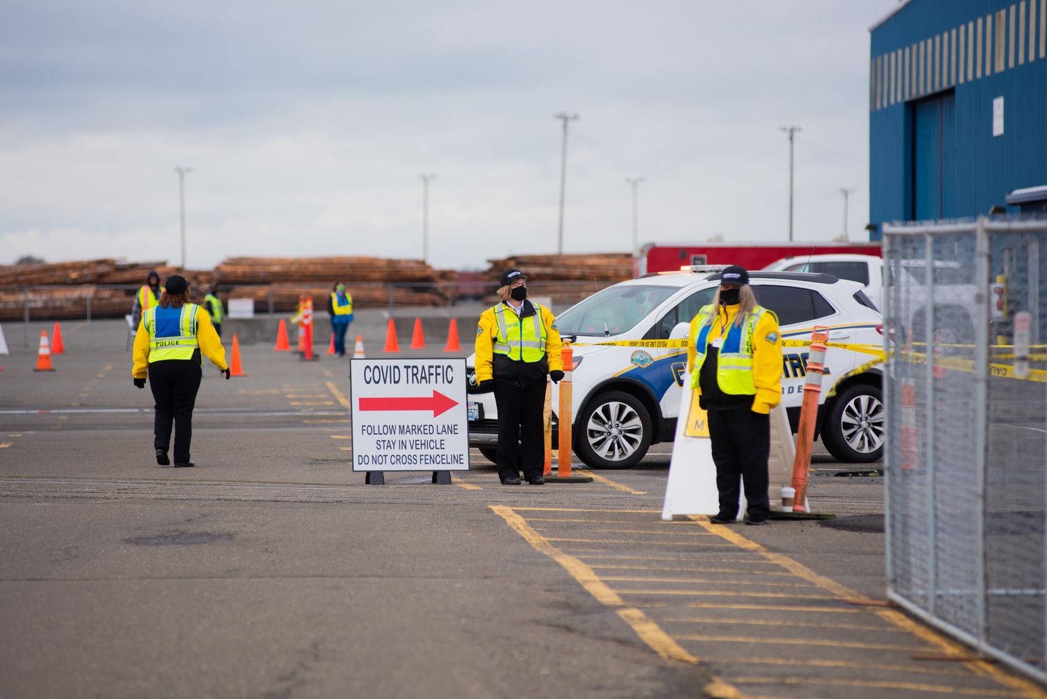 Traffic officers in yellow high-visibility vests direct vehicles beside a police car and a road sign in a parking lot with orange cones.