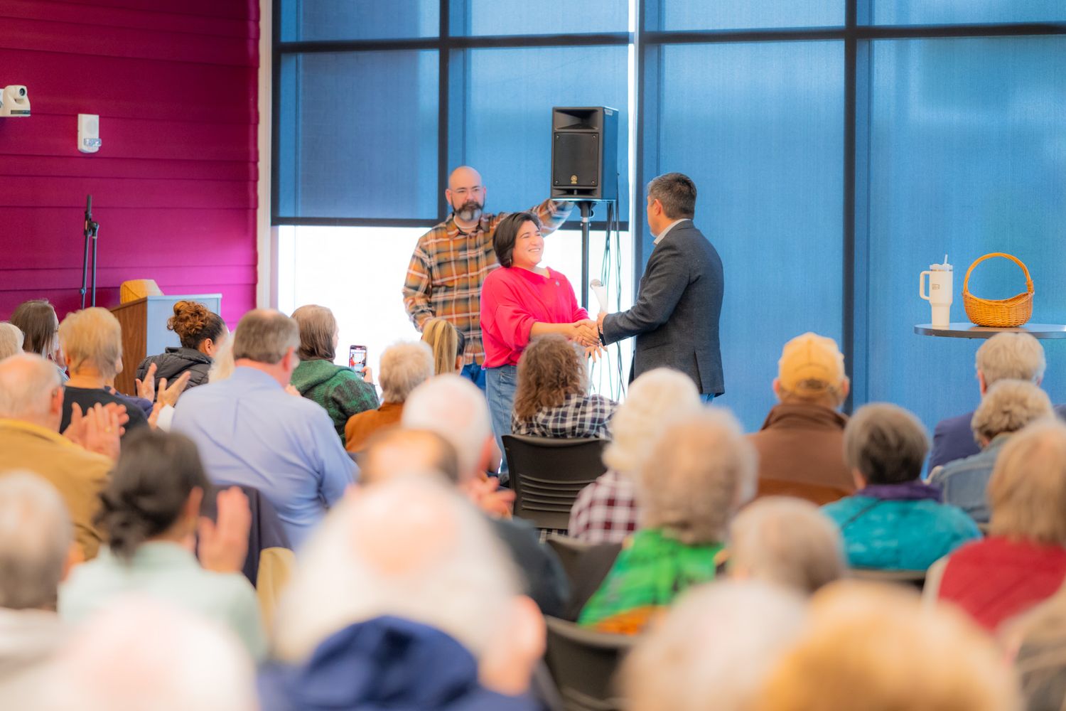 A woman in a red top addresses a seated audience of older adults while two men stand nearby in a bright room with large windows.