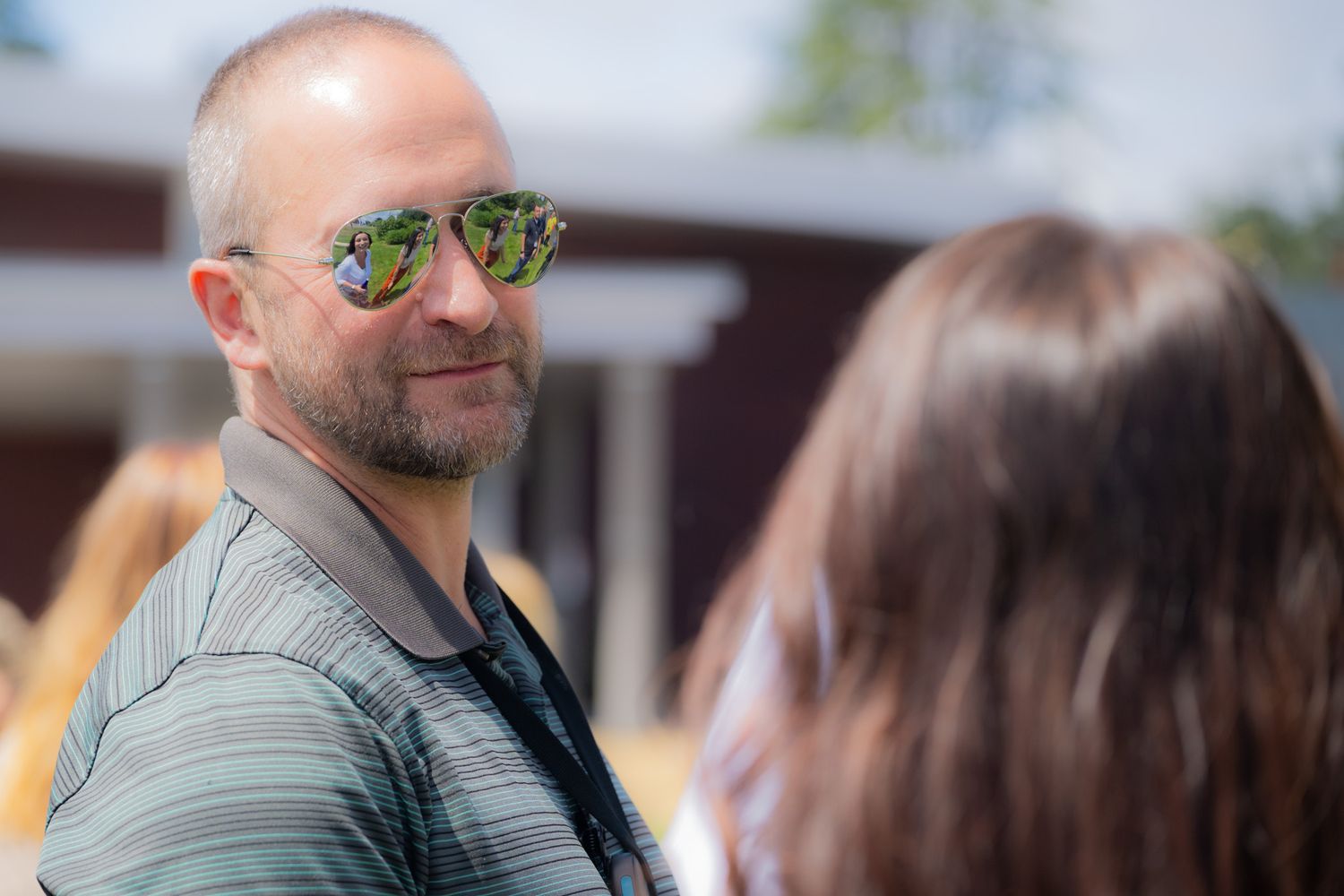 Smiling bald man with a goatee wearing mirrored aviator sunglasses and a striped polo shirt talks with a long-haired person outdoors.