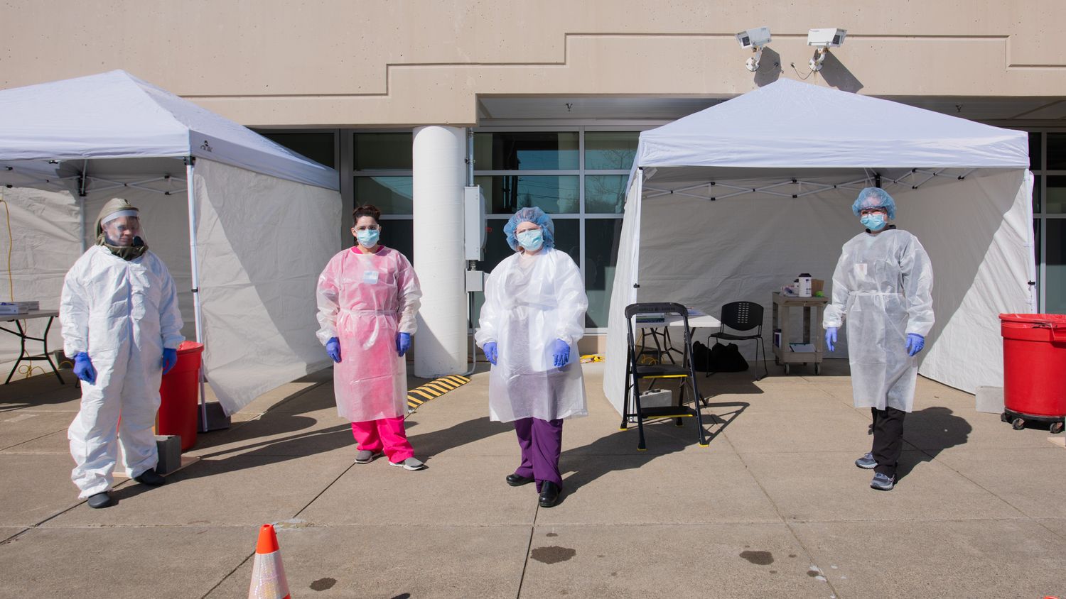 Four healthcare workers in protective gowns, masks and face shields stand outside white medical tents at an outdoor testing site.