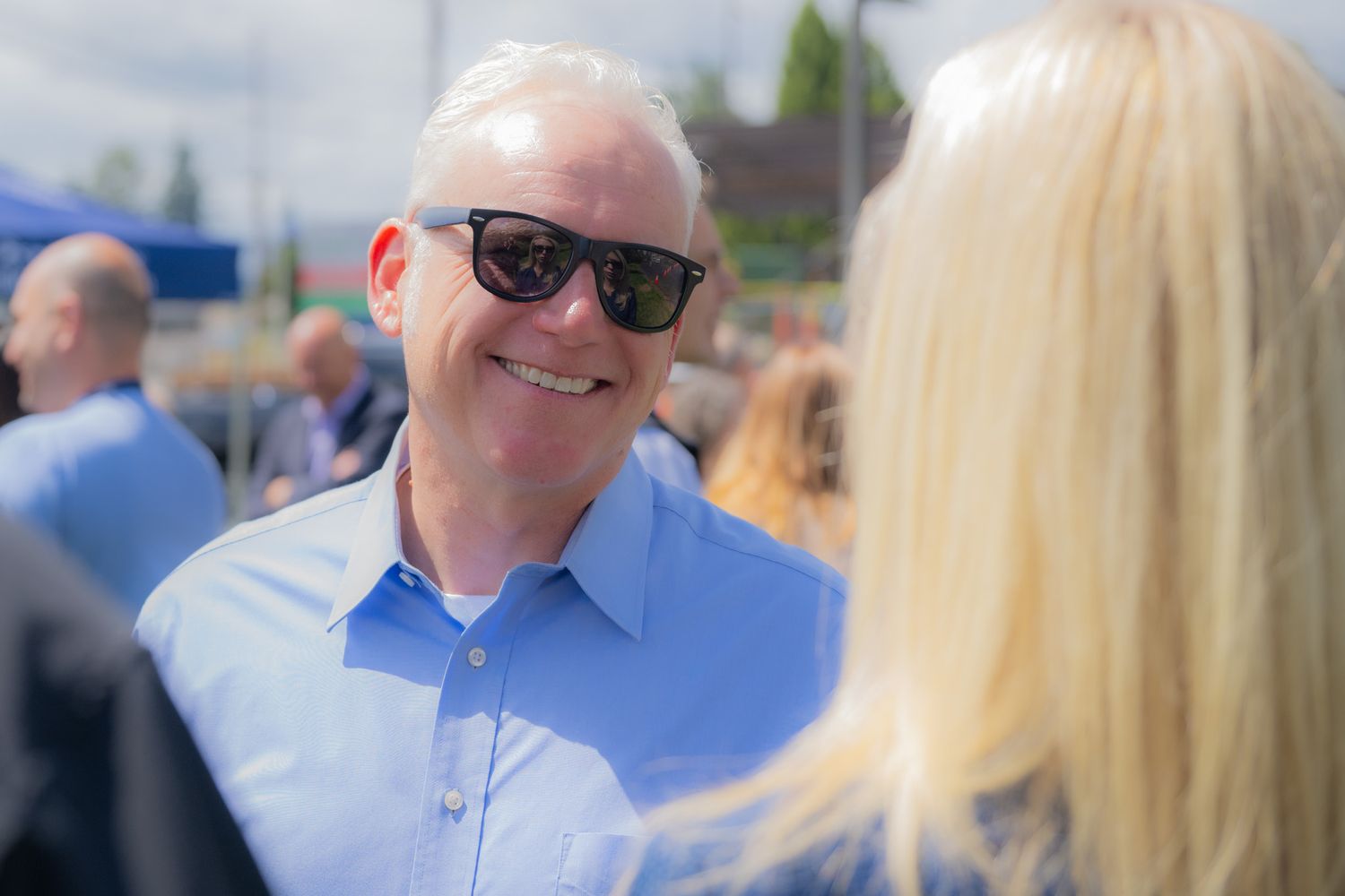 Smiling man with white hair wearing sunglasses and a light blue button-up shirt talks with a blonde woman at a sunny outdoor gathering.