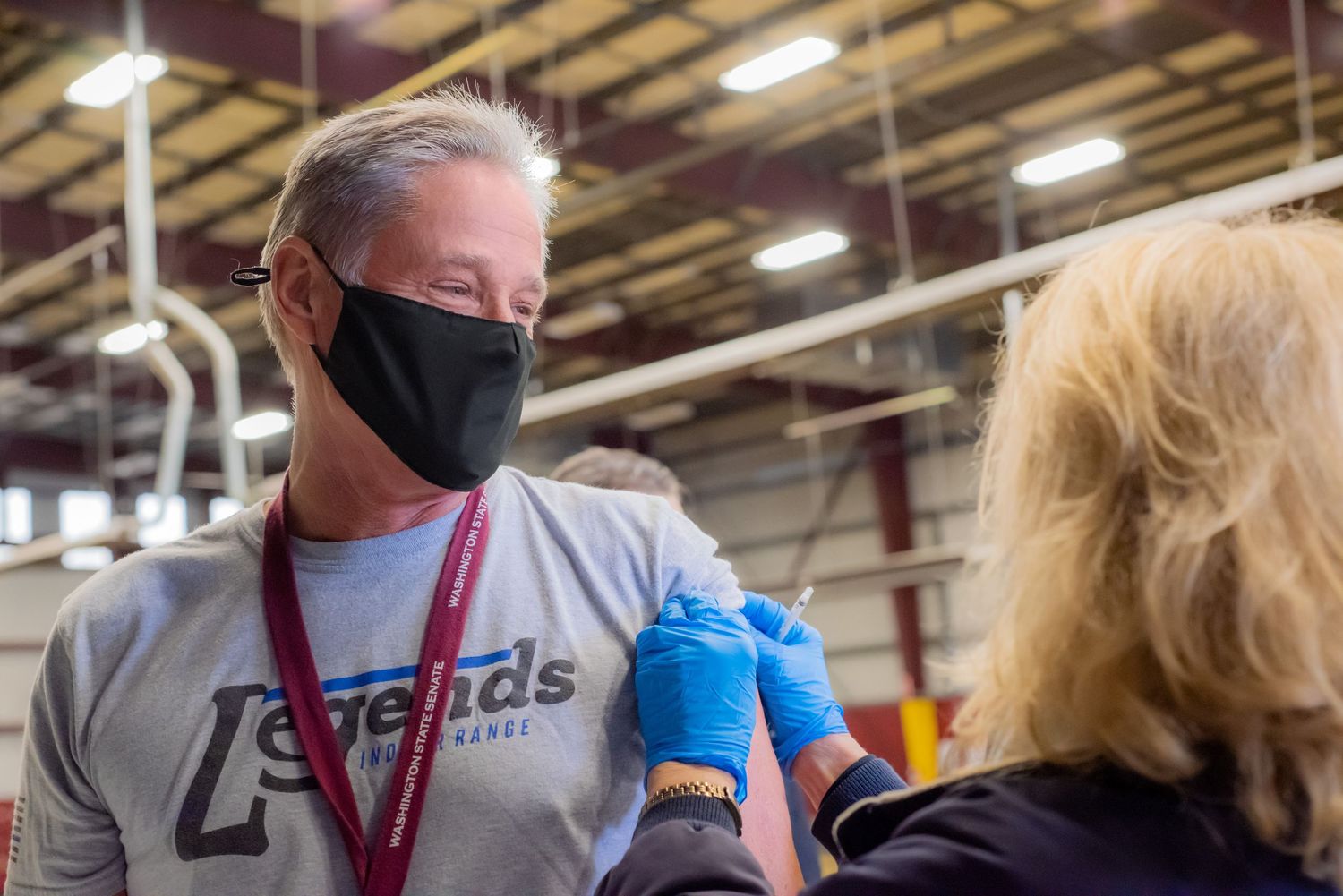 Man in a black face mask and gray Legends t-shirt receives a vaccine injection from a gloved healthcare worker at an indoor clinic.