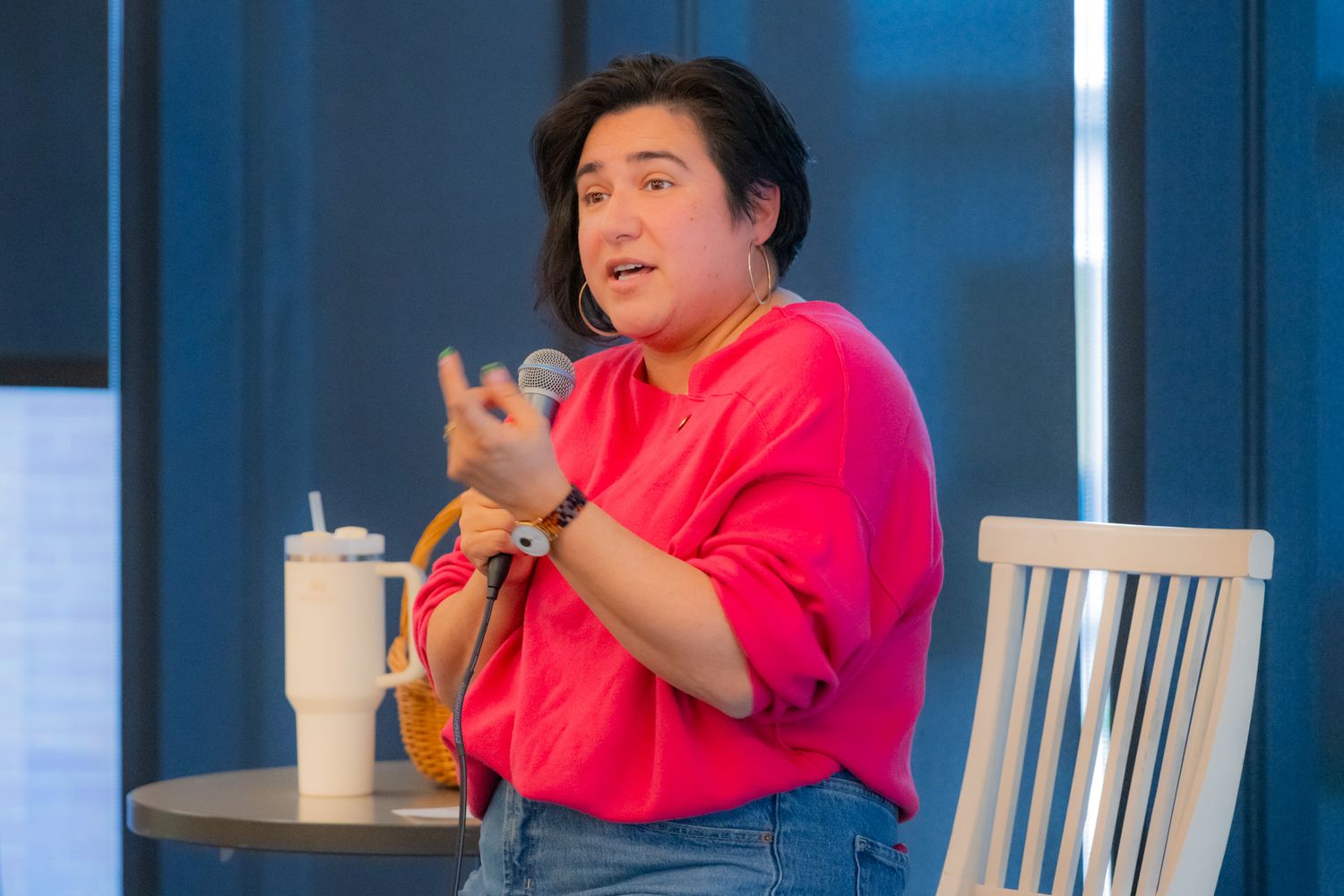 A woman in a pink sweater speaks into a microphone beside a small table holding a tumbler and a wicker basket.