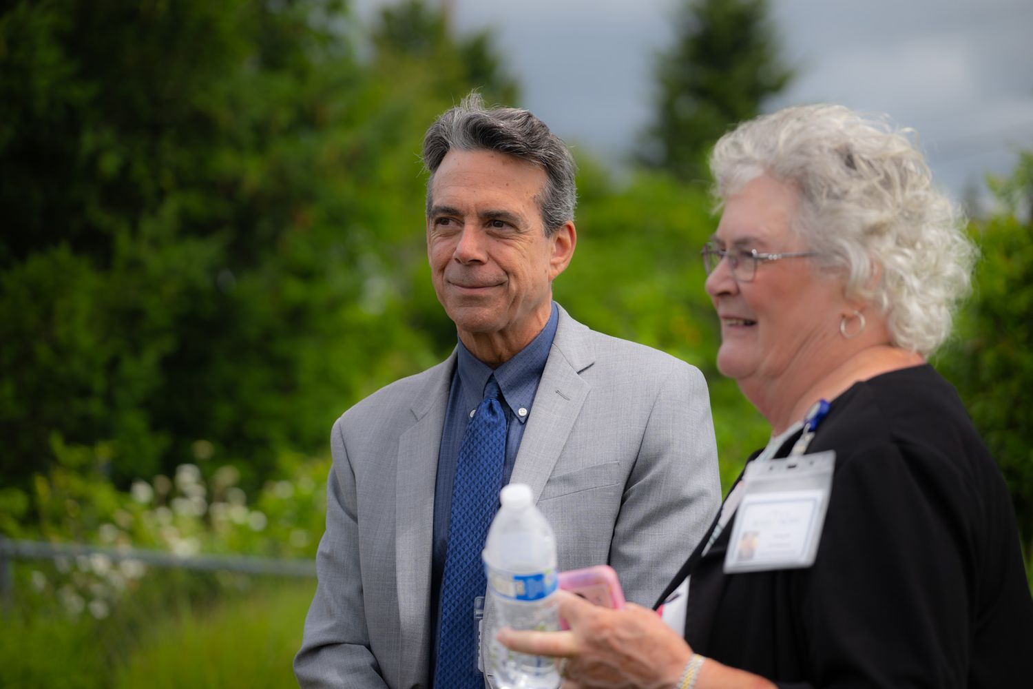 A gray-haired man in a gray blazer and blue shirt stands beside a smiling older woman wearing a name tag, holding a water bottle outdoors.