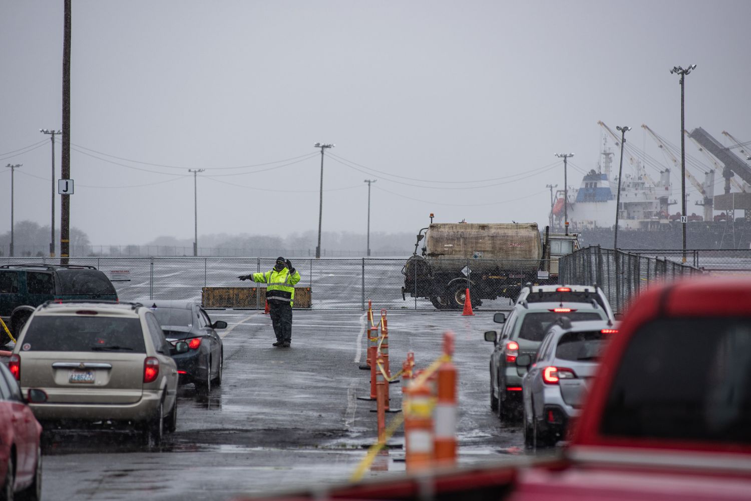 Worker in a high-visibility jacket directs lines of cars at a wet port checkpoint with cranes and a cargo ship in the background.