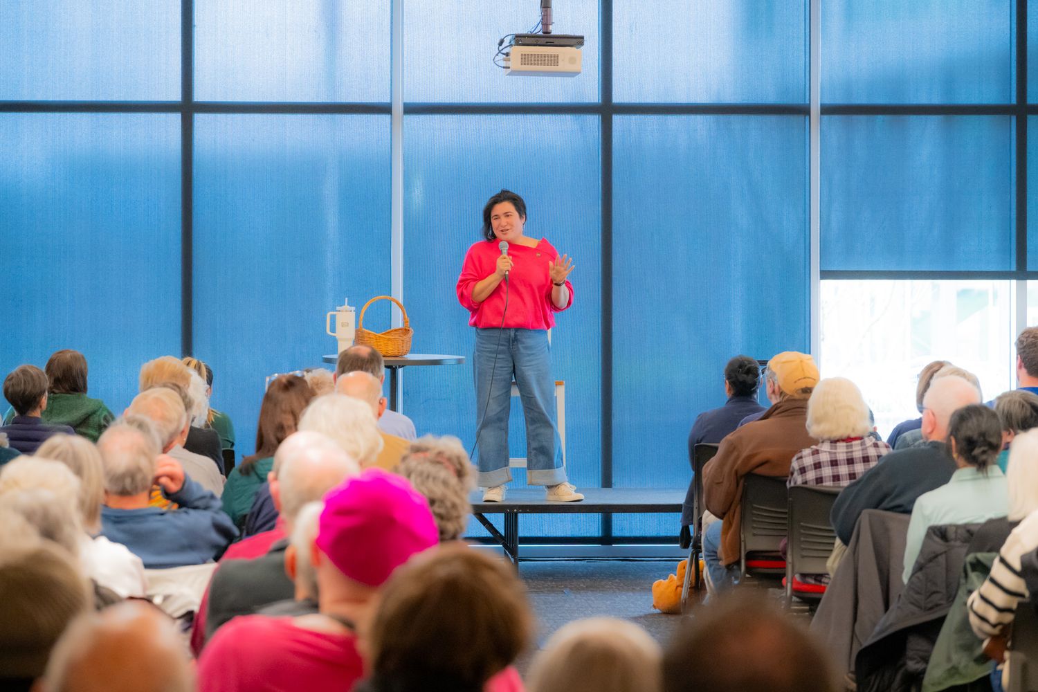 A woman in a bright pink top and jeans speaks to a seated audience in a large room with tall blue windows during a community gathering.