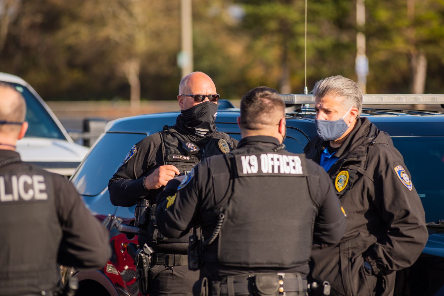 Uniformed police officers wearing face masks talk beside a patrol vehicle in a parking lot.