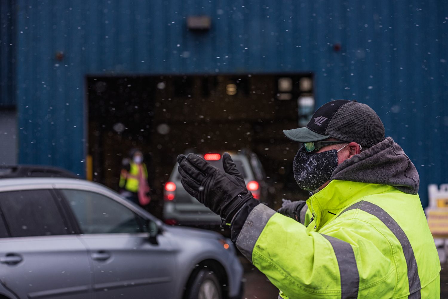 A masked worker in a high-visibility yellow jacket and cap gestures to direct a vehicle at a drive-through site in falling snow.