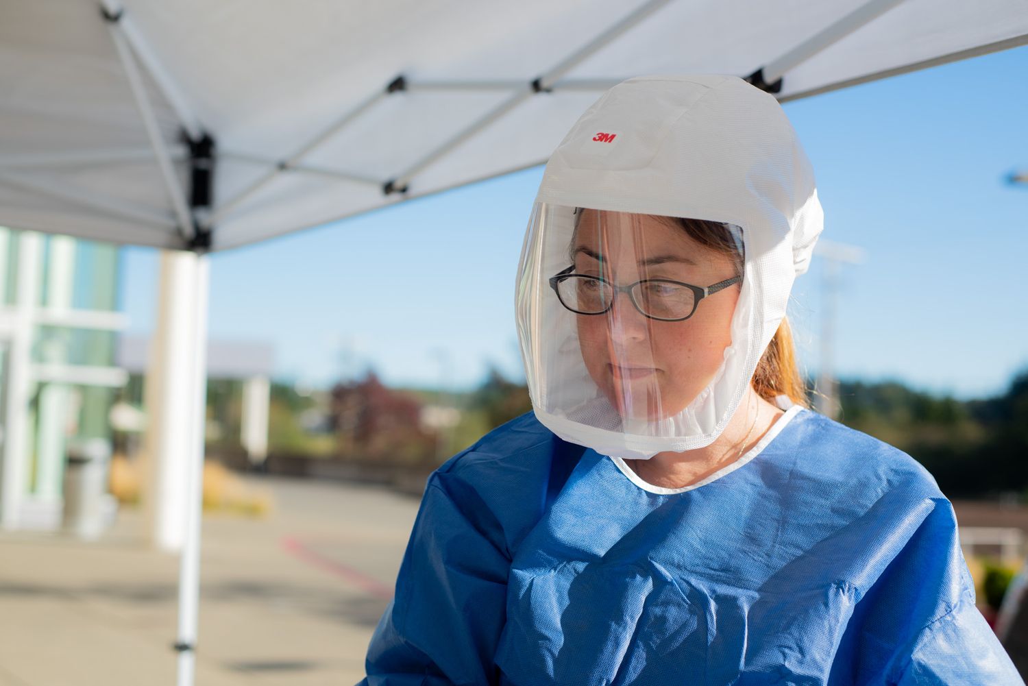 Healthcare worker in a blue protective gown, face shield and hood works outdoors under a white tent at a medical testing site.