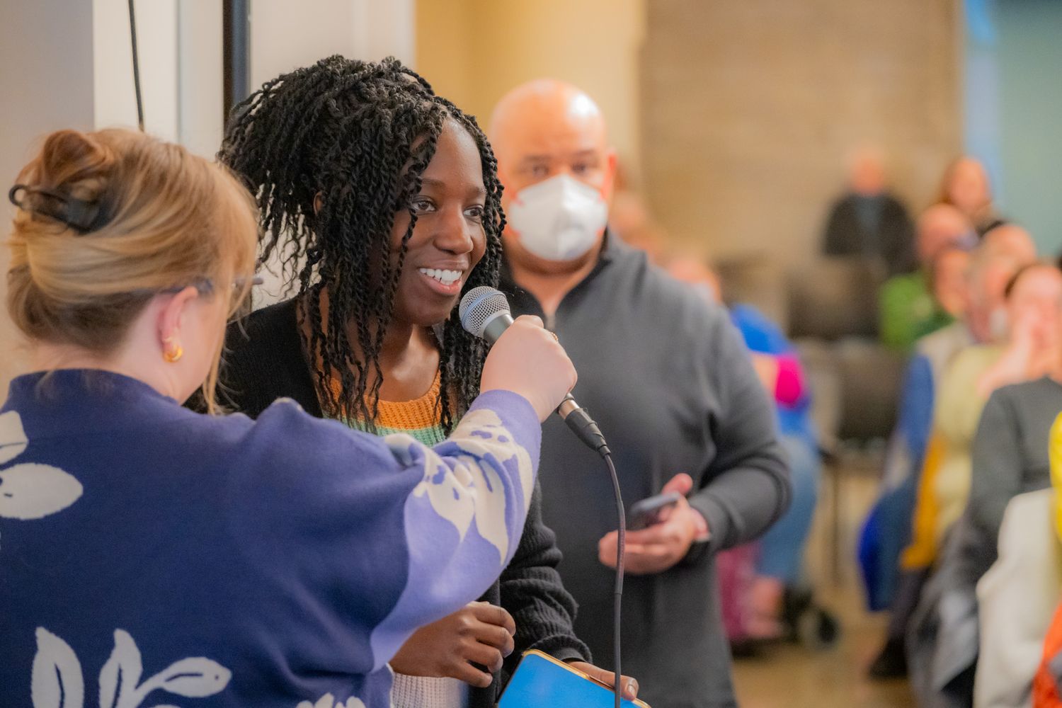 A woman with long braids smiles while speaking into a microphone held by another woman as a masked man and audience look on.