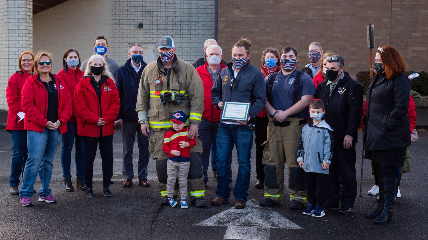 Masked community members in red jackets, a firefighter, and a child pose outdoors while holding a framed certificate in a parking lot.