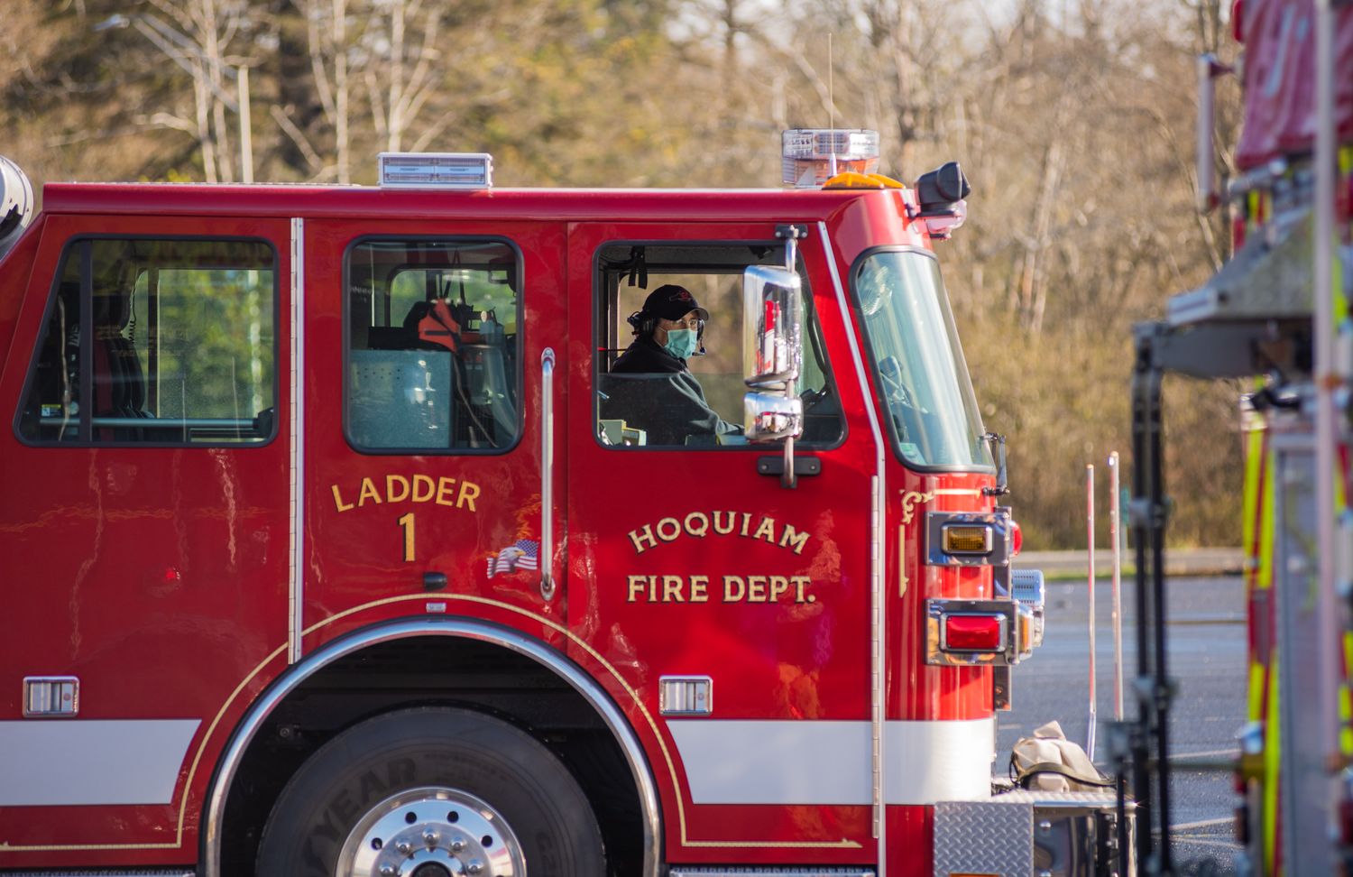 Red Hoquiam Fire Department ladder truck with a masked firefighter in the cab drives past on a sunny day.