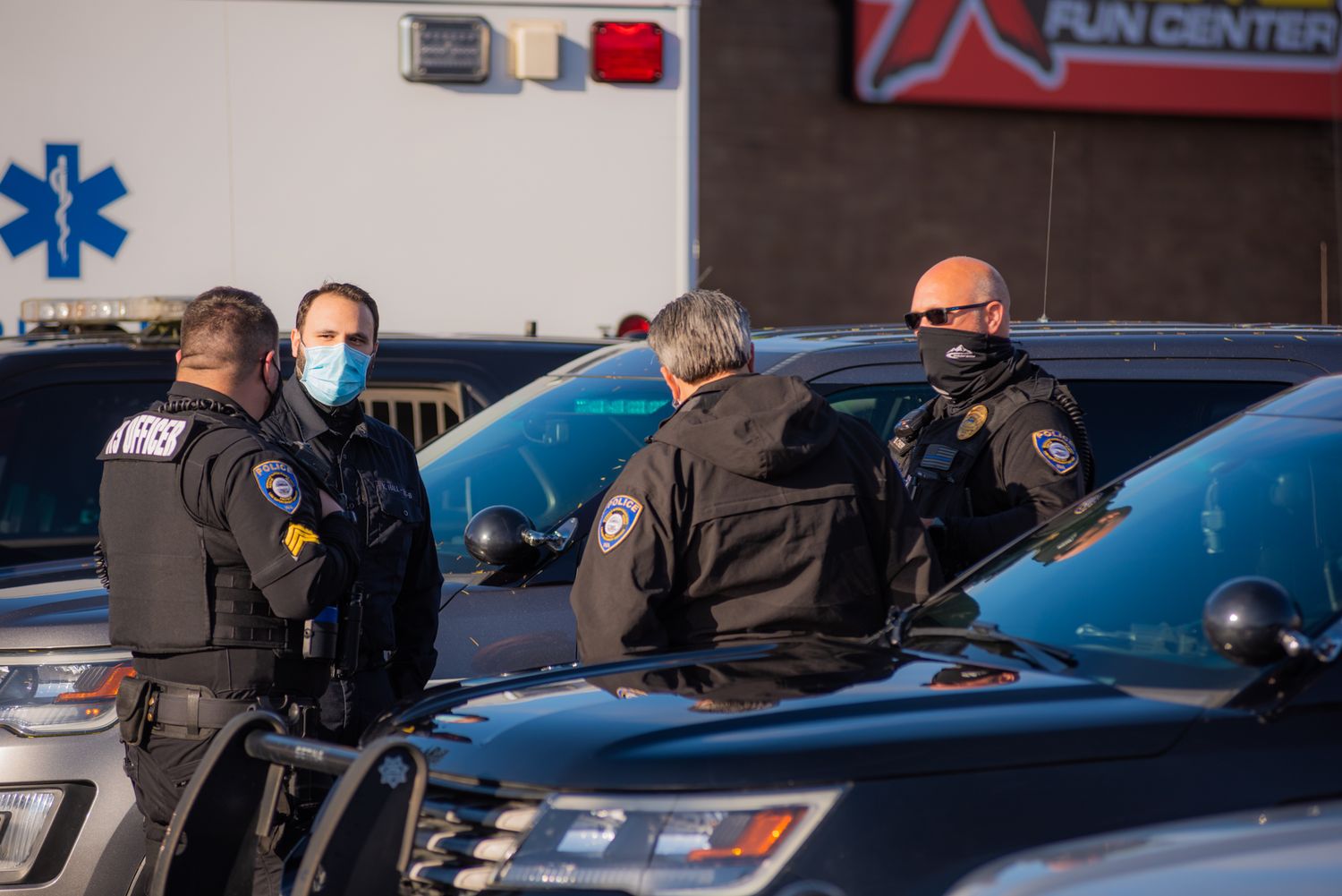 Masked police officers and first responders gather around patrol cars in front of an ambulance in a parking lot.
