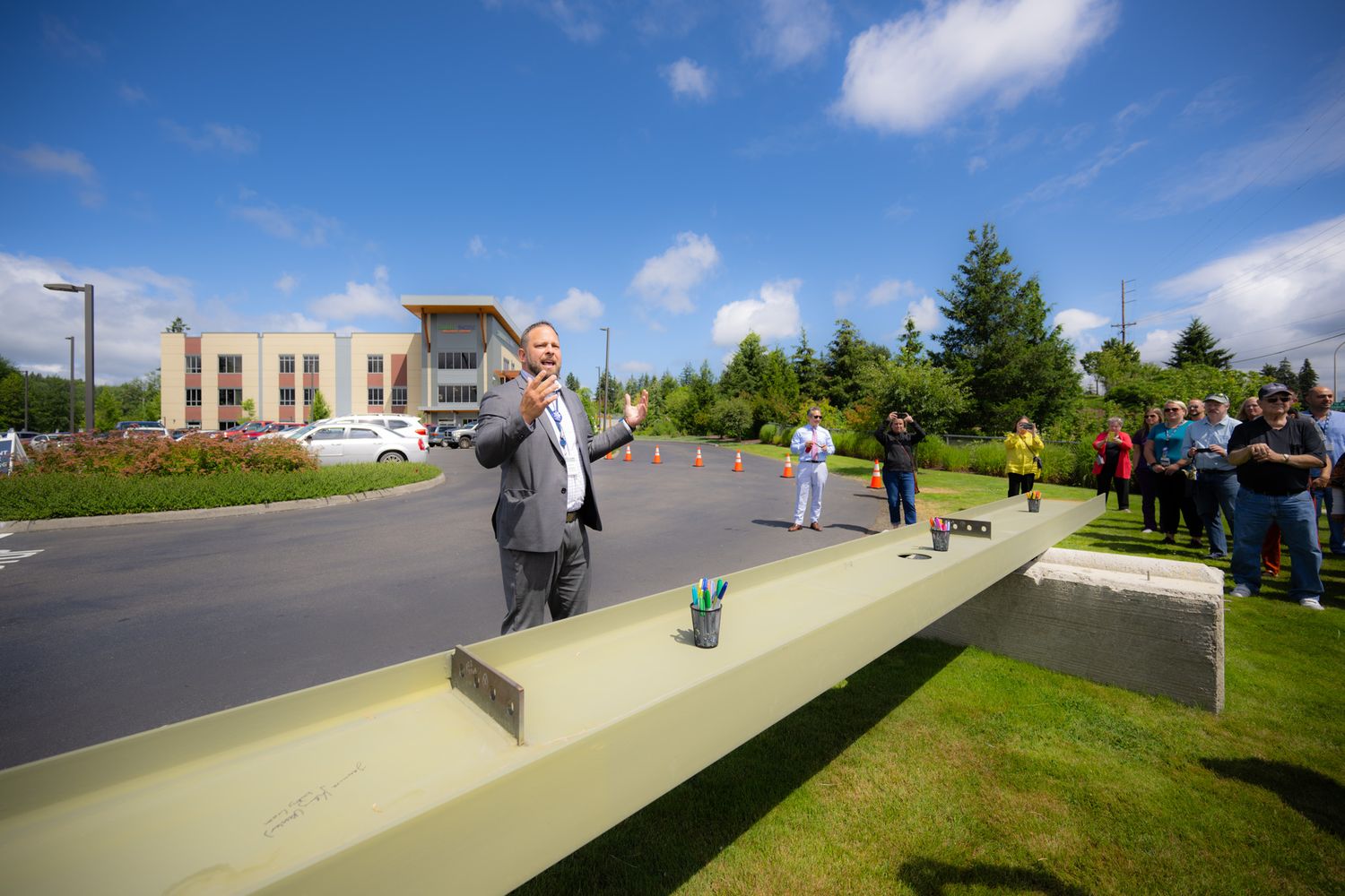 Man in a gray suit speaks into a microphone beside a large steel beam as onlookers gather on the lawn near a modern office building.