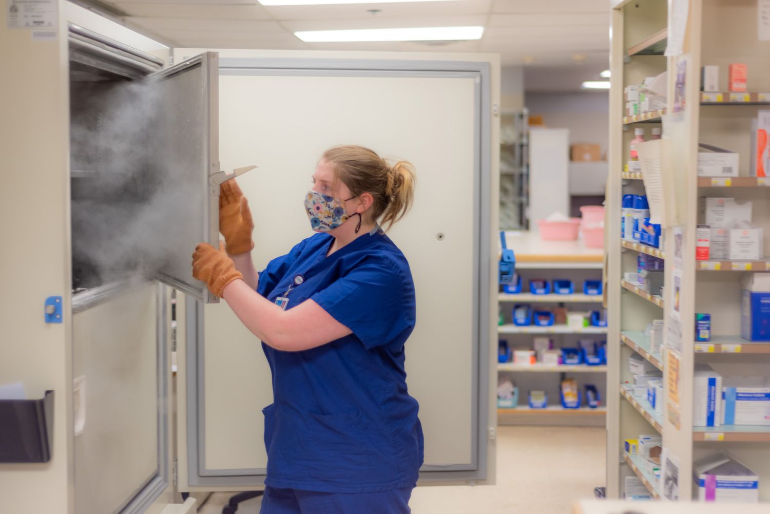 A healthcare worker in blue scrubs, mask and gloves opens an ultra-cold freezer releasing vapor in a pharmacy storage room.