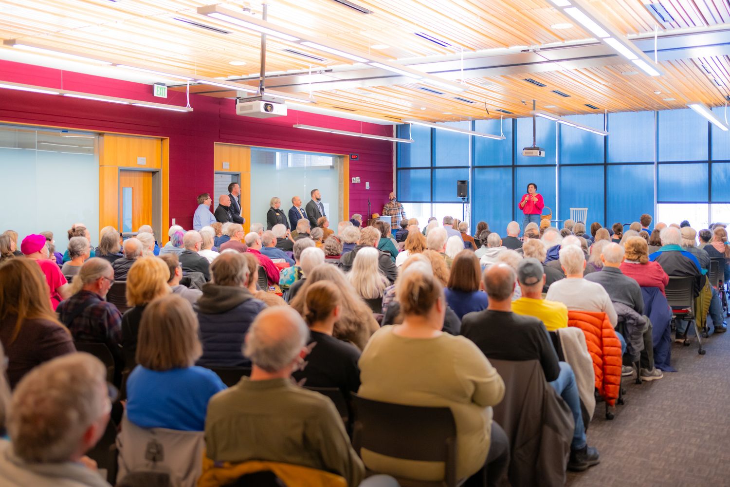 A large audience of seated attendees listens to a speaker presenting at the front of a bright, modern auditorium with red walls.