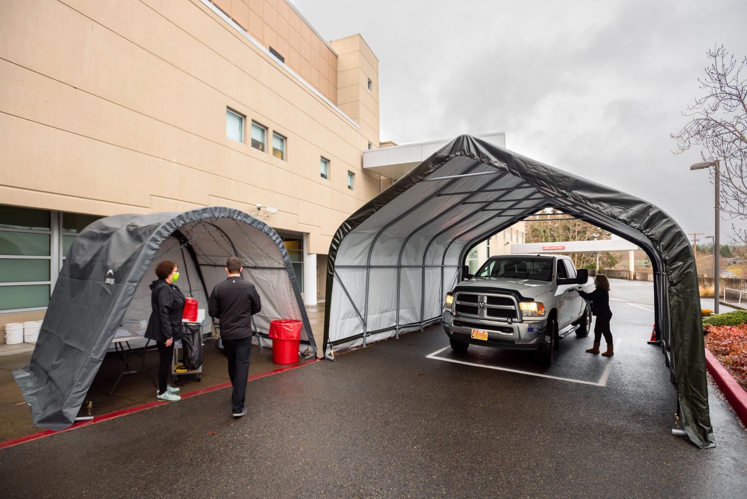 Pickup truck drives into a temporary tent canopy outside a hospital building while two people in protective gear stand near a second tent.