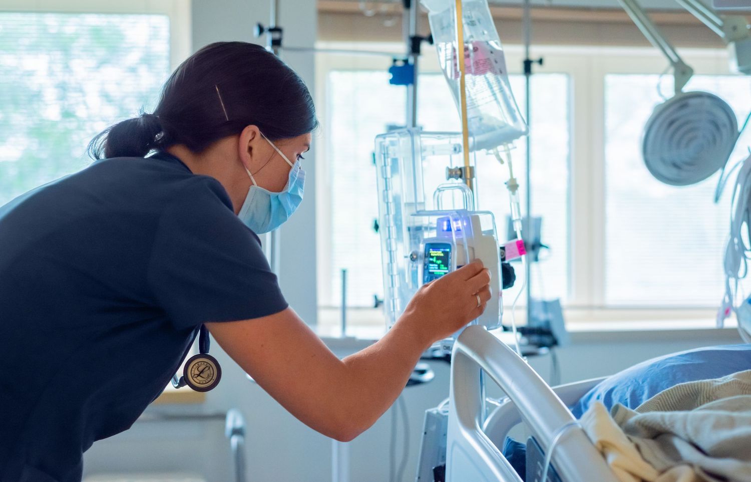 A masked nurse in dark scrubs adjusts an IV infusion pump beside a hospital bed in a bright patient room.