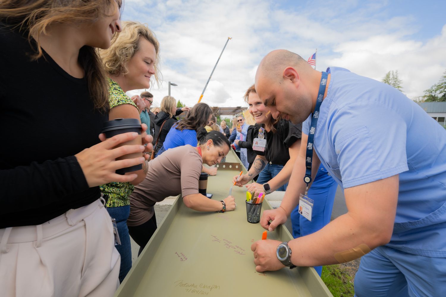 Group of people in casual clothes gather around a long outdoor table, leaning in to play a game together on a cloudy day.