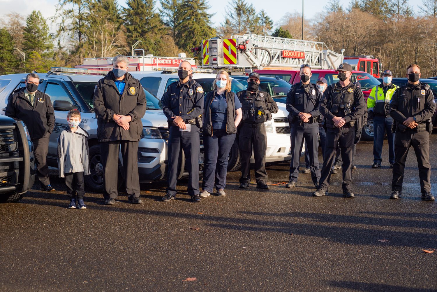 A line of masked police and emergency responders stand in a parking lot with a small child in front and fire trucks behind.
