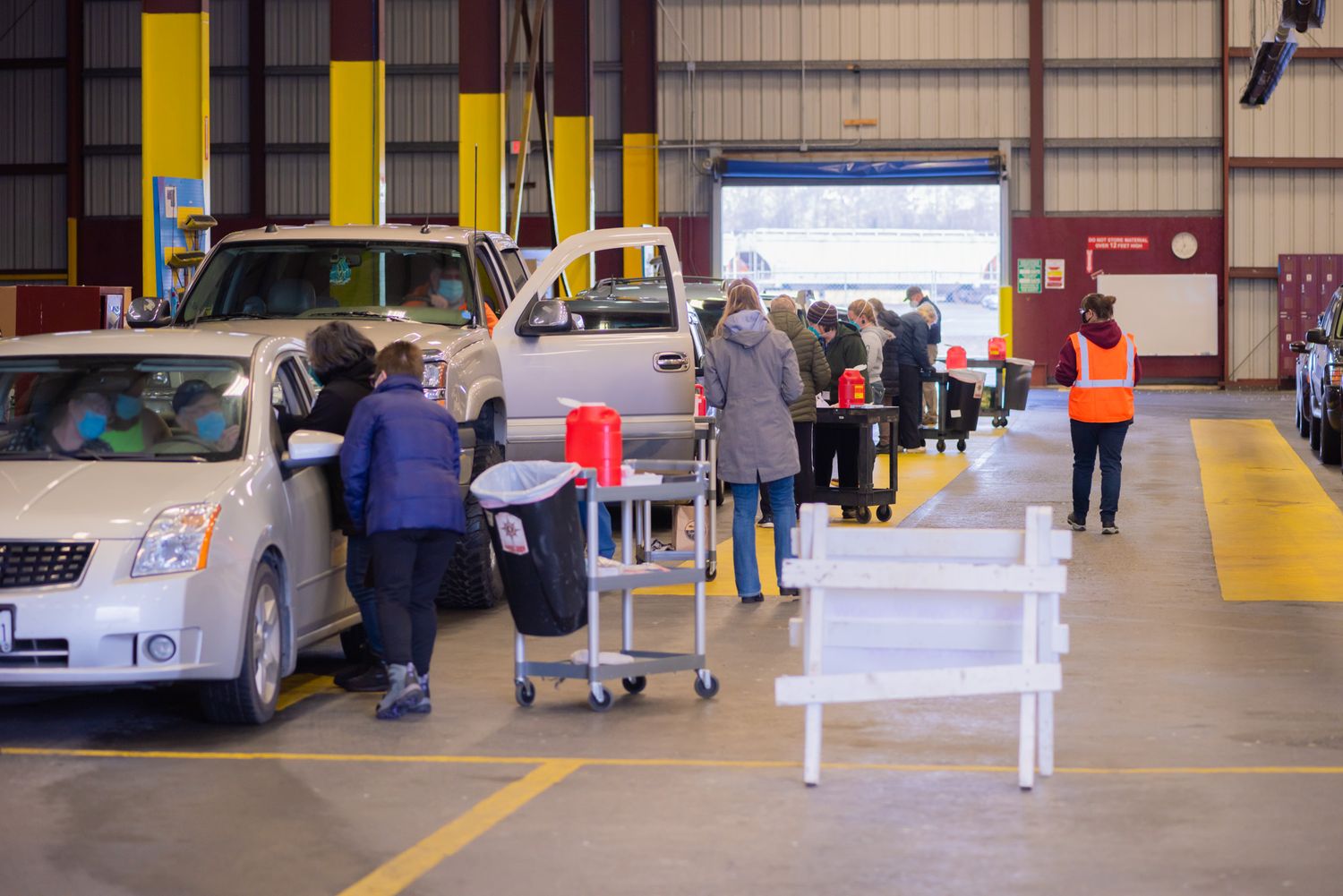 Staff in safety vests assist people beside parked cars at an indoor drive-through vaccination site inside a large warehouse.