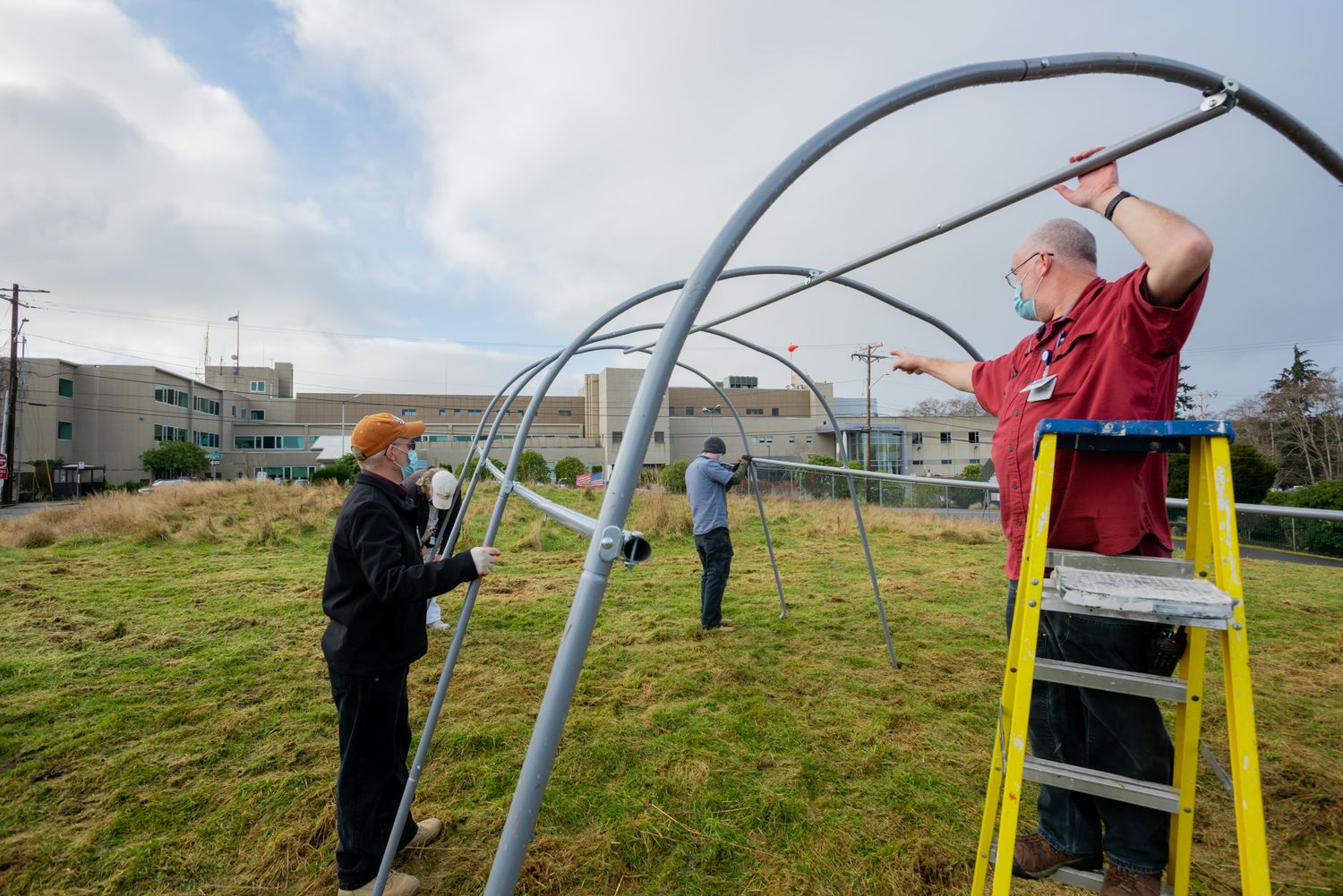 Volunteers on a grassy field assemble a metal hoop house greenhouse frame, one man on a yellow ladder, with buildings in the background.