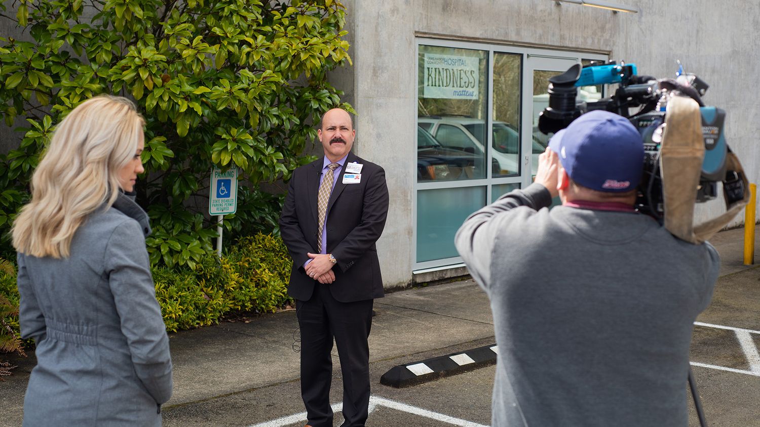 A man in a dark suit stands outdoors facing a camera operator filming him, while a blonde woman watches nearby in front of a building.