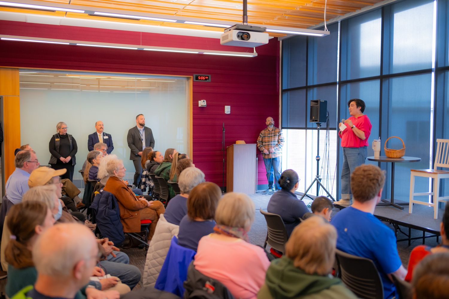 Seated audience members listen to a woman speaking at a community event in a bright room with large windows and a projector overhead.