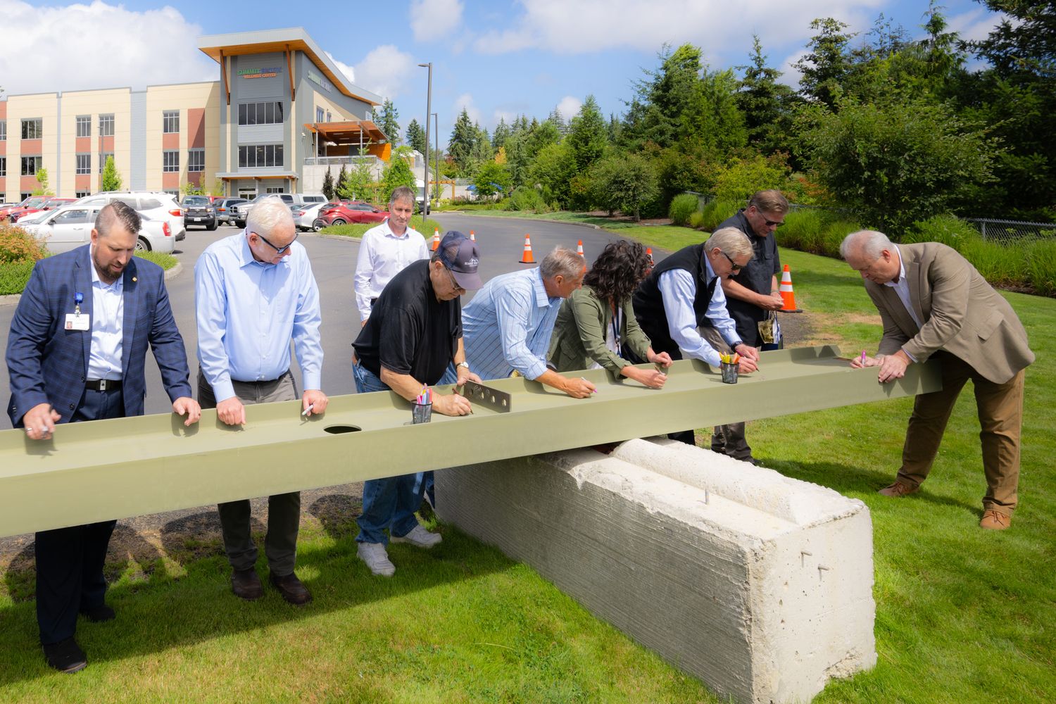 Group of men in business casual attire signing a long green steel beam outdoors on a lawn during a building construction ceremony.