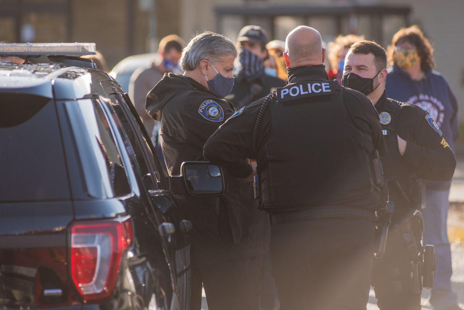 Police officers wearing face masks stand in a group beside a parked patrol vehicle during an outdoor gathering.
